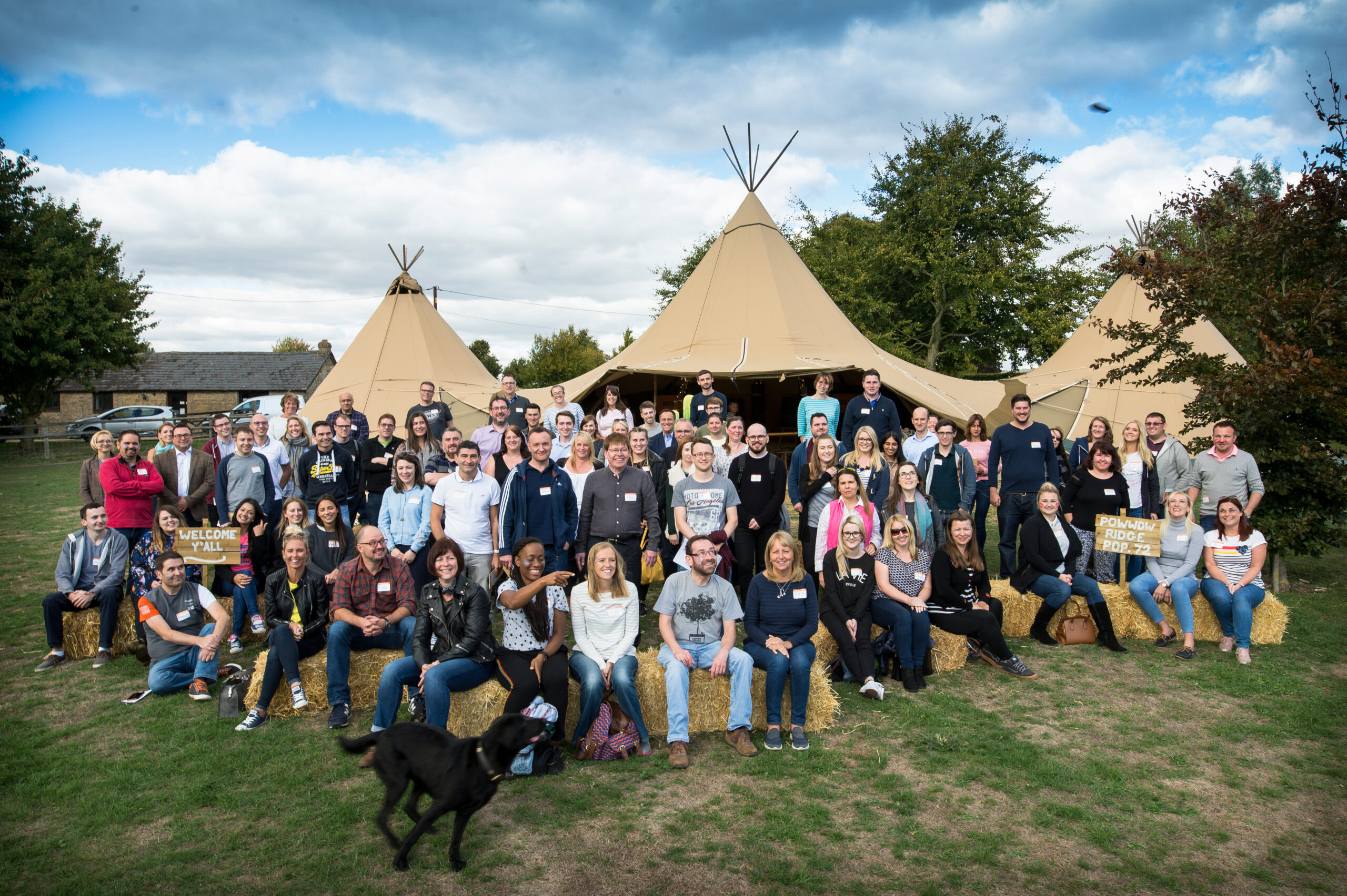 A group of about 80 people are gathered in front of three teepees. The front front row is seated on hay bales. The rest stand. They are facing the camera.