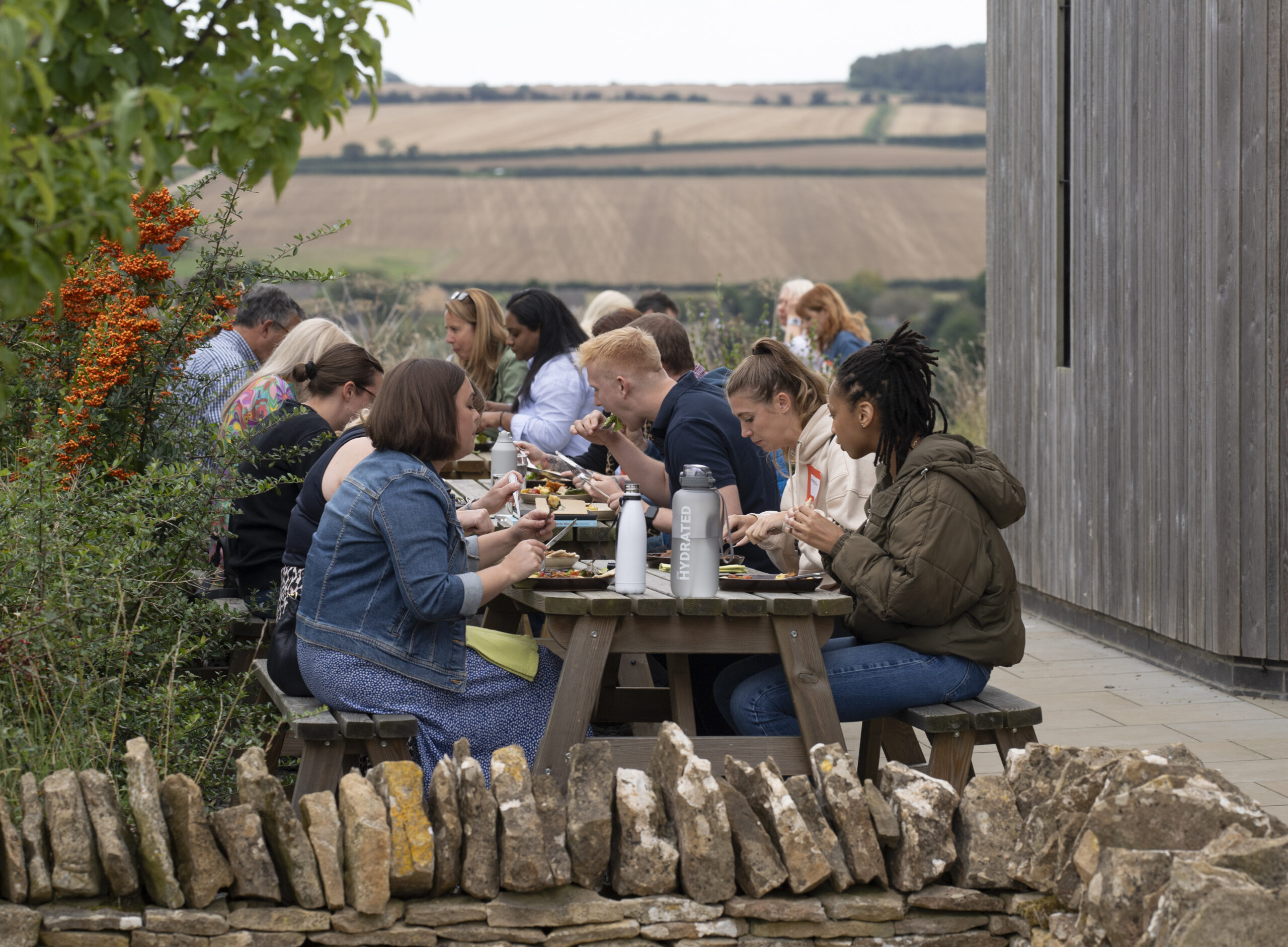 Paraplanner share a meal at a wooden picnic benches outdoors in a rural setting. The tables are filled with food and drinks. Behind the diners, farmland stretches to the horizon. In the foreground, a stone wall adds rustic charm. Foliage frames the scene, including vivid orange berries.