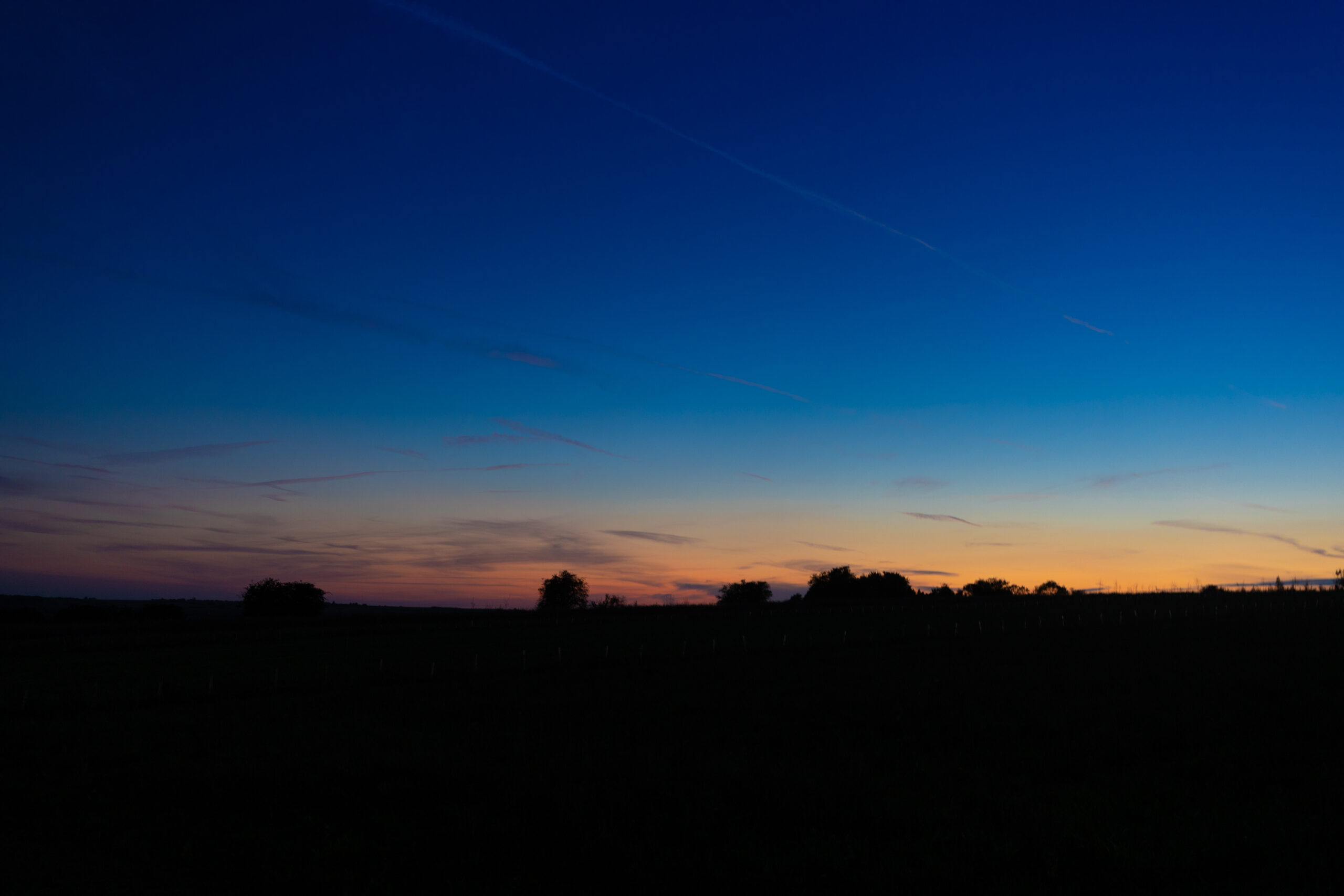 A serene rural sunset landscape. The sky dominates, showcasing a gradient from deep blue at the top to warm orange and pink near the horizon. Thin, wispy clouds are scattered across the sky. The lower third is a dark silhouette of flat countryside with a few trees and bushes visible along the horizon. The foreground is nearly black, suggesting an open field. The contrast between the vibrant sky and dark landscape creates a striking composition.