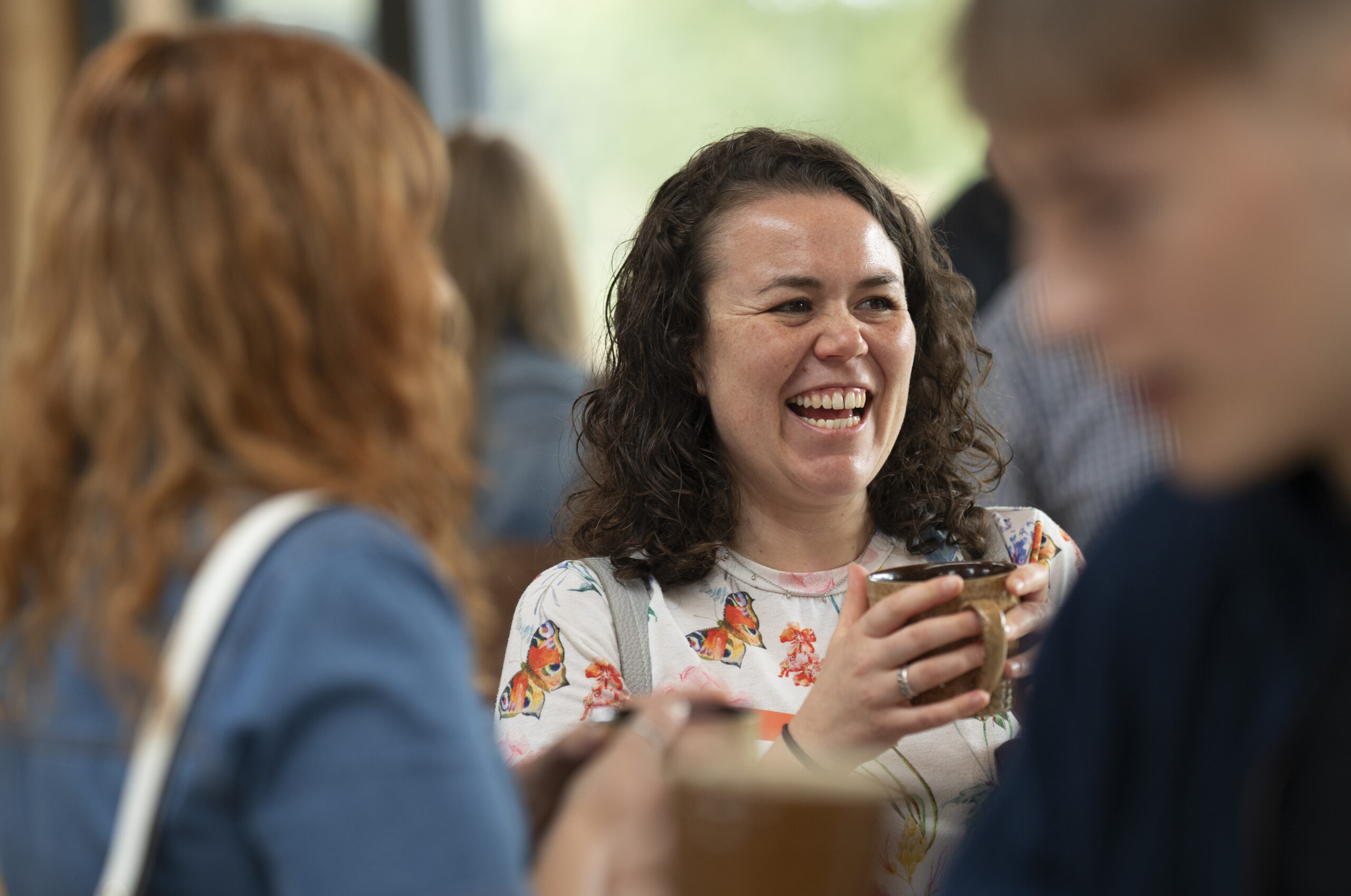 A woman with curly dark hair is laughing.Her face expresses genuine joy. She's wearing a white top with colourful butterfly prints and holding a mug. She appears to be in conversation with others, visible partially in the foreground.