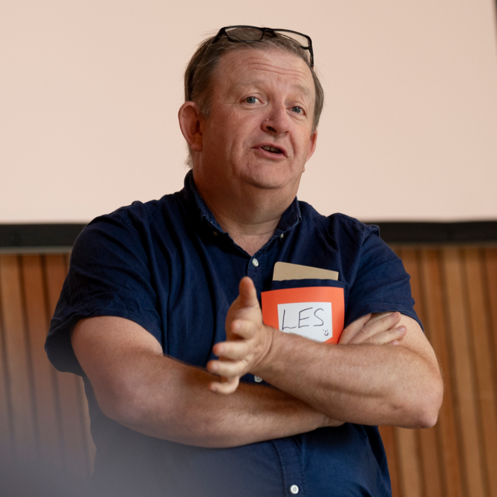 A waist-up shot of Les Cameron speaking at The Big Day Out event in 2023. He is dressed informally in a dark blue short-sleeved shirt. His arms are folded across his chest. His glasses are perched on top of his head as he looks towards us and lightly upwards as though thinking while he talks.