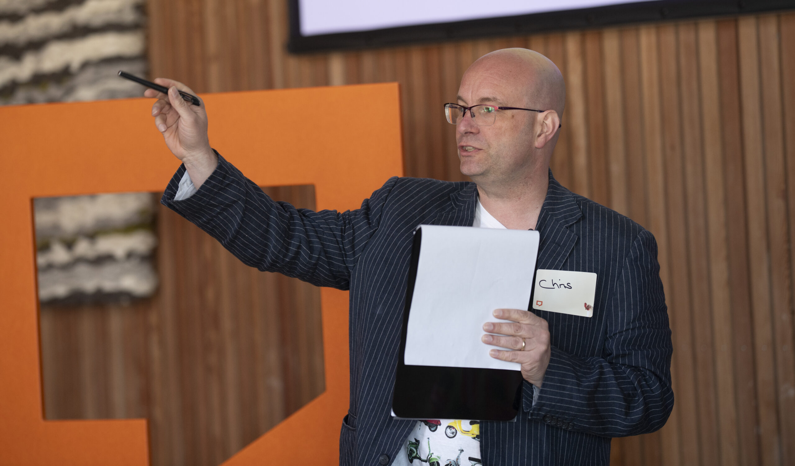 Chris Wormwell explains the schedule for the day during the opening session of The Big Day Out 2024. He's standing and holds a clipboard in his left hand. He is pointing with his right hand and his arm outstretched. He's standing in front of a wooden vertically- panelled wall. To Chris’s right, part of the Paraplanners’ Assembly logo is visible.