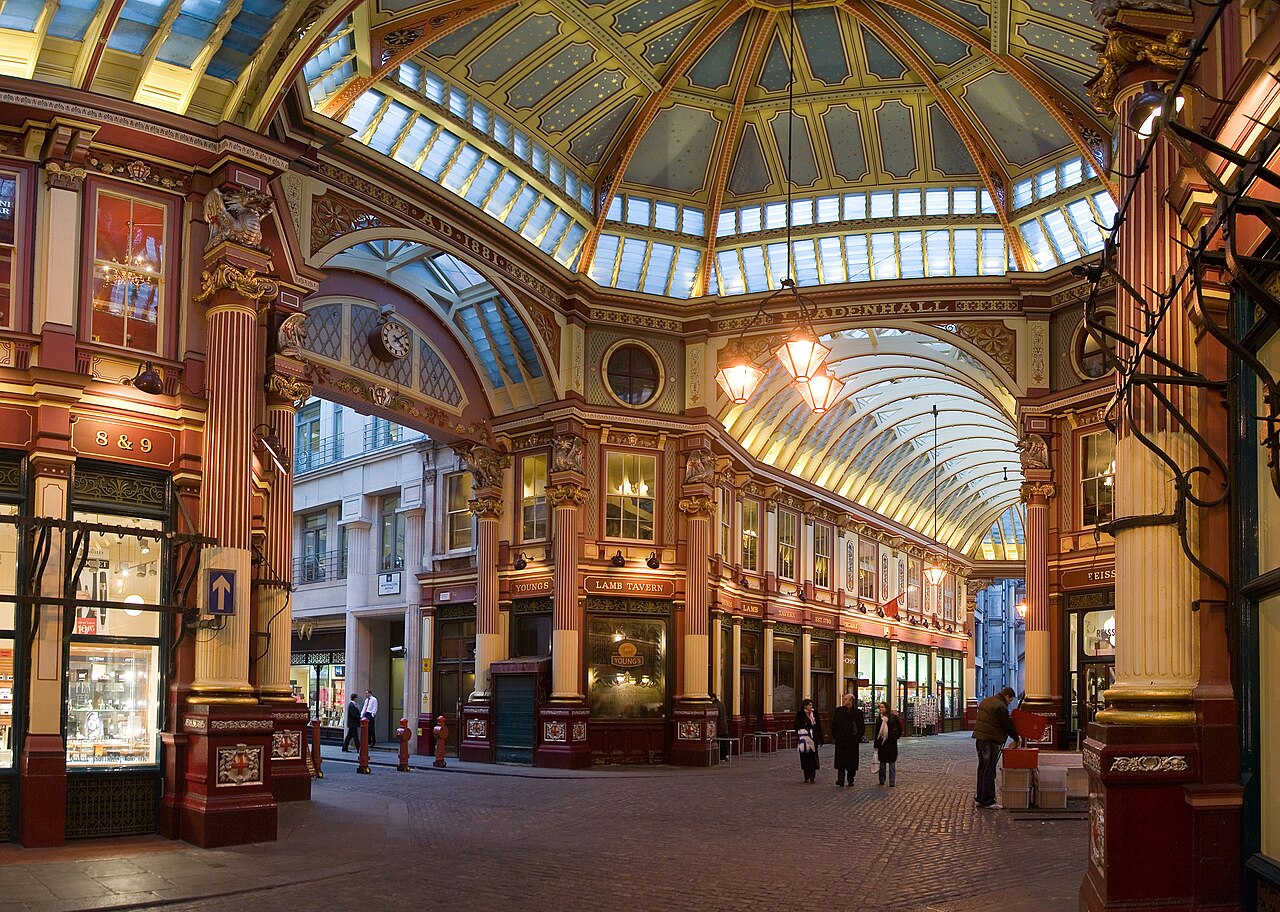 The image shows an ornate Victorian-style indoor shopping arcade with a grand glass ceiling. Elaborate architectural details include gilded arches, columns, and decorative mouldings. The space features storefronts with illuminated displays, including a pub called "Lamb Tavern". A few people can be seen walking through the elegantly lit passage. 