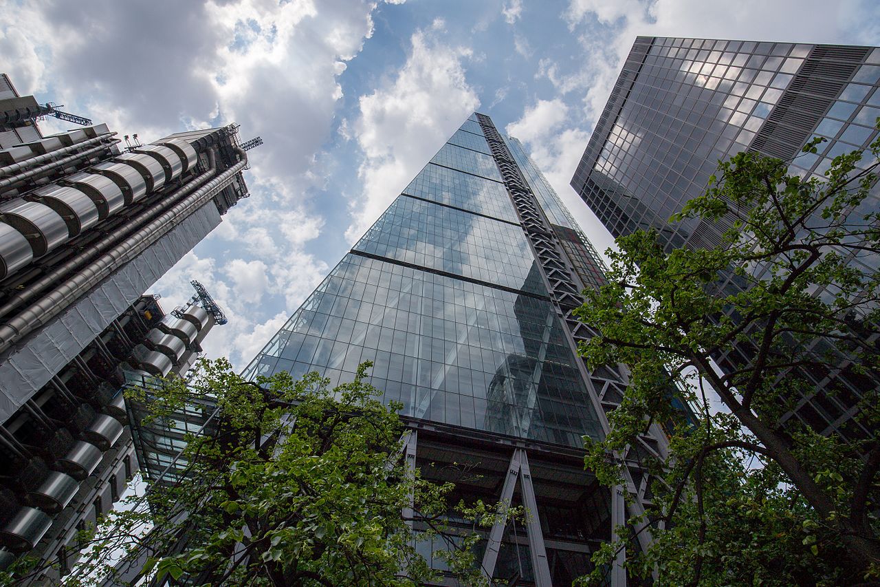 A horizontal low-angle view of The Leadenhall Building in the City of London. The building's distinctive 'cheese grater' shape is nestled next to the Lloyd's building on the left, with its exposed metallic structure, and sleek glass-fronted towers in the center and right. Green tree branches frame the bottom of the image, while a partly cloudy blue sky fills the background above.