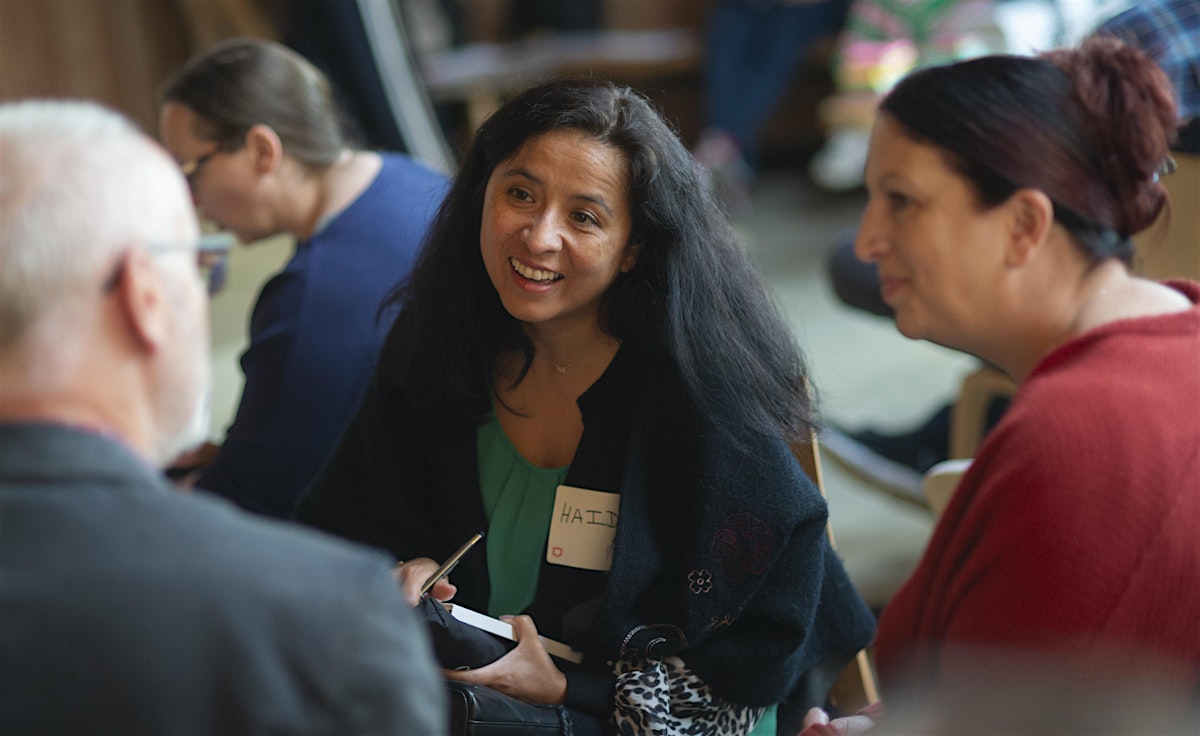 In the centre of the image sits a smiling woman with long dark hair. She's wearing a green top and dark jacket. She's engaged in conversation with others around her, including a person in a red shirt visible to the right of the image. The picture is from The Big Day Out 2024.