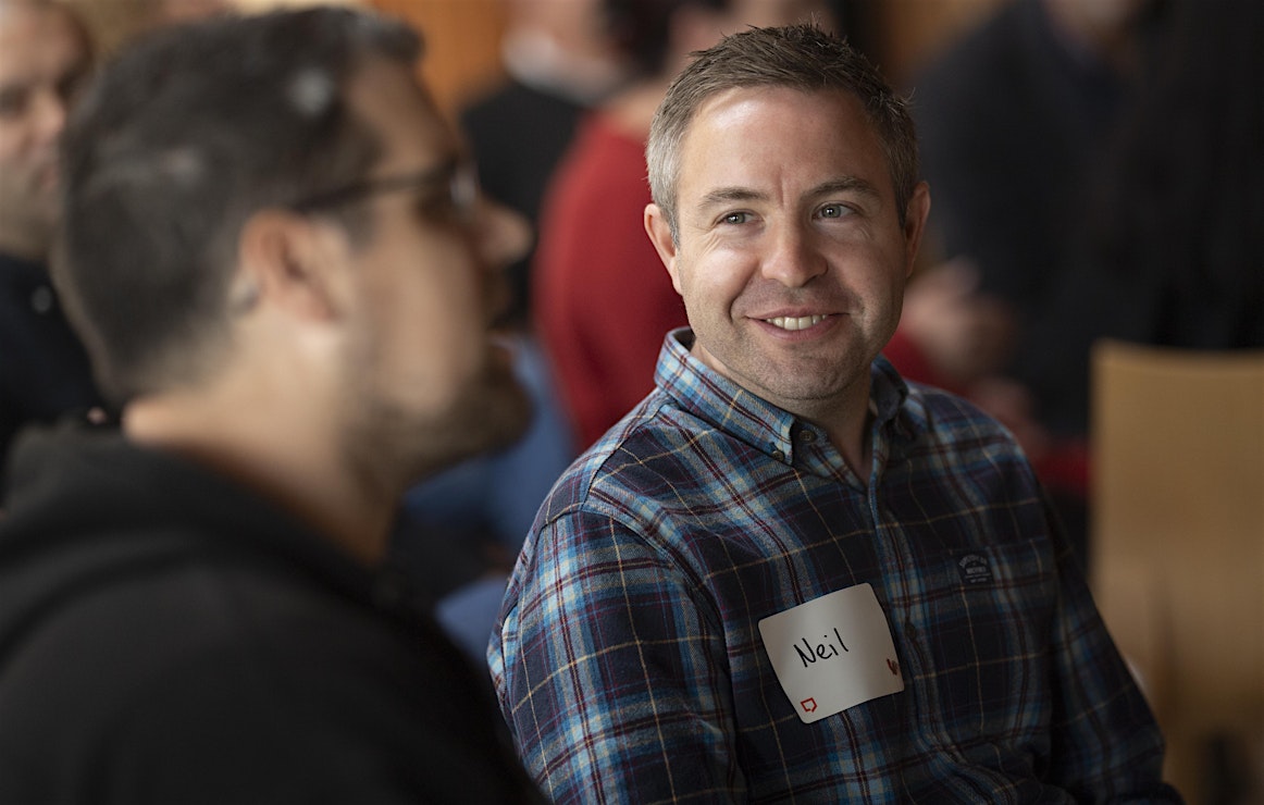 The focus of the image is a person in a blue tartan checked shirt who is smiling. His handwritten name badge says 'Neil'. He appears to be listening to a person who is pictured nearest to the camera but out of focus. The image was taken at the Paraplanners' Assembly's Big Day Out 2024.