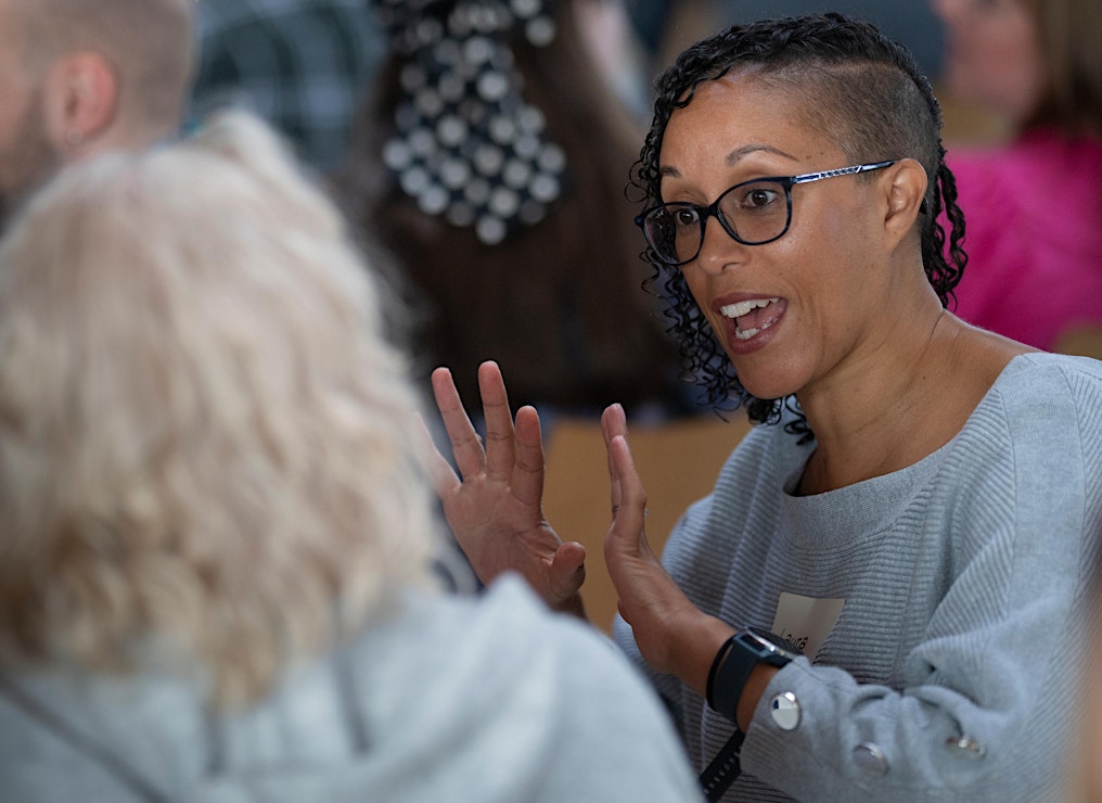 The focus of the image is a person gesturing expressively with their hands while speaking animatedly. They are in conversation with another person who is out of focus in the foreground on the left-hand side of the image. 